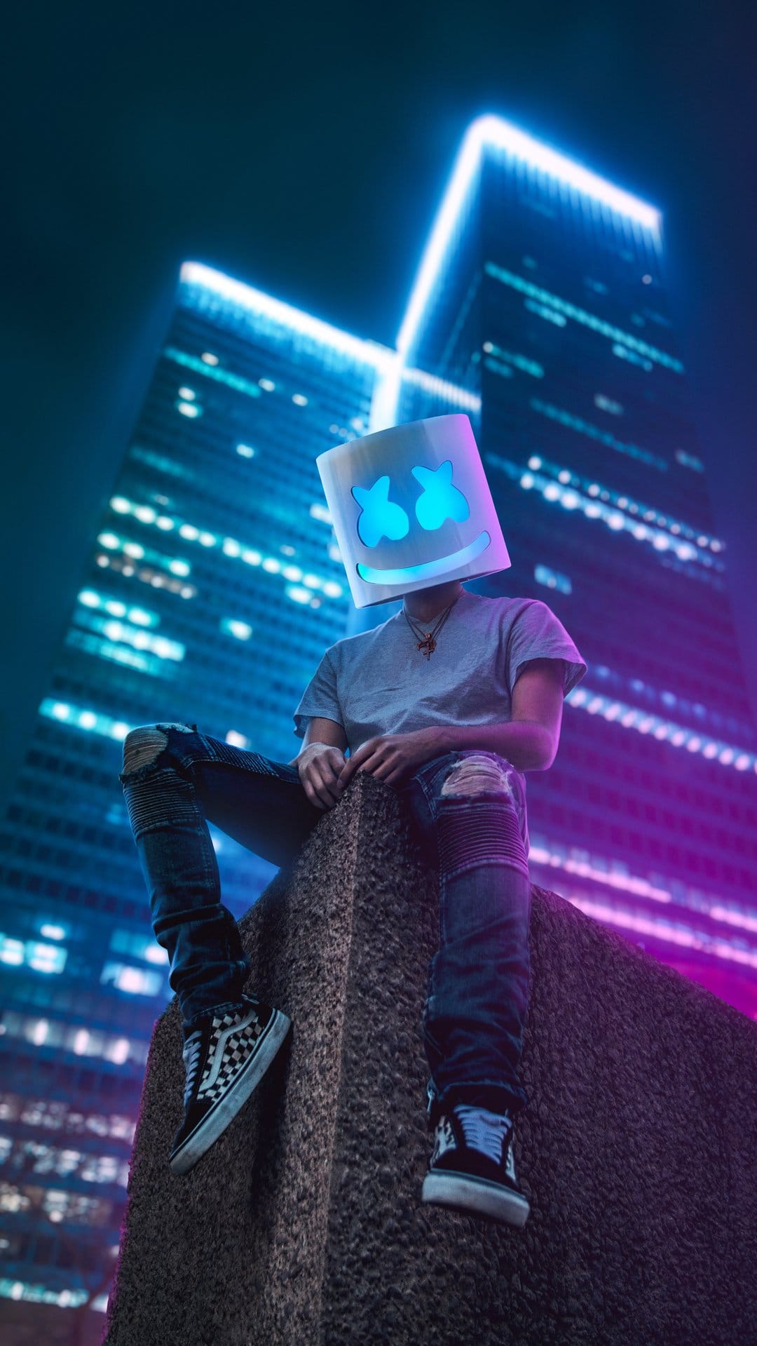 Marshmello in a white bucket helmet sits on a stone ledge before glowing blue and purple neon skyscrapers at night.