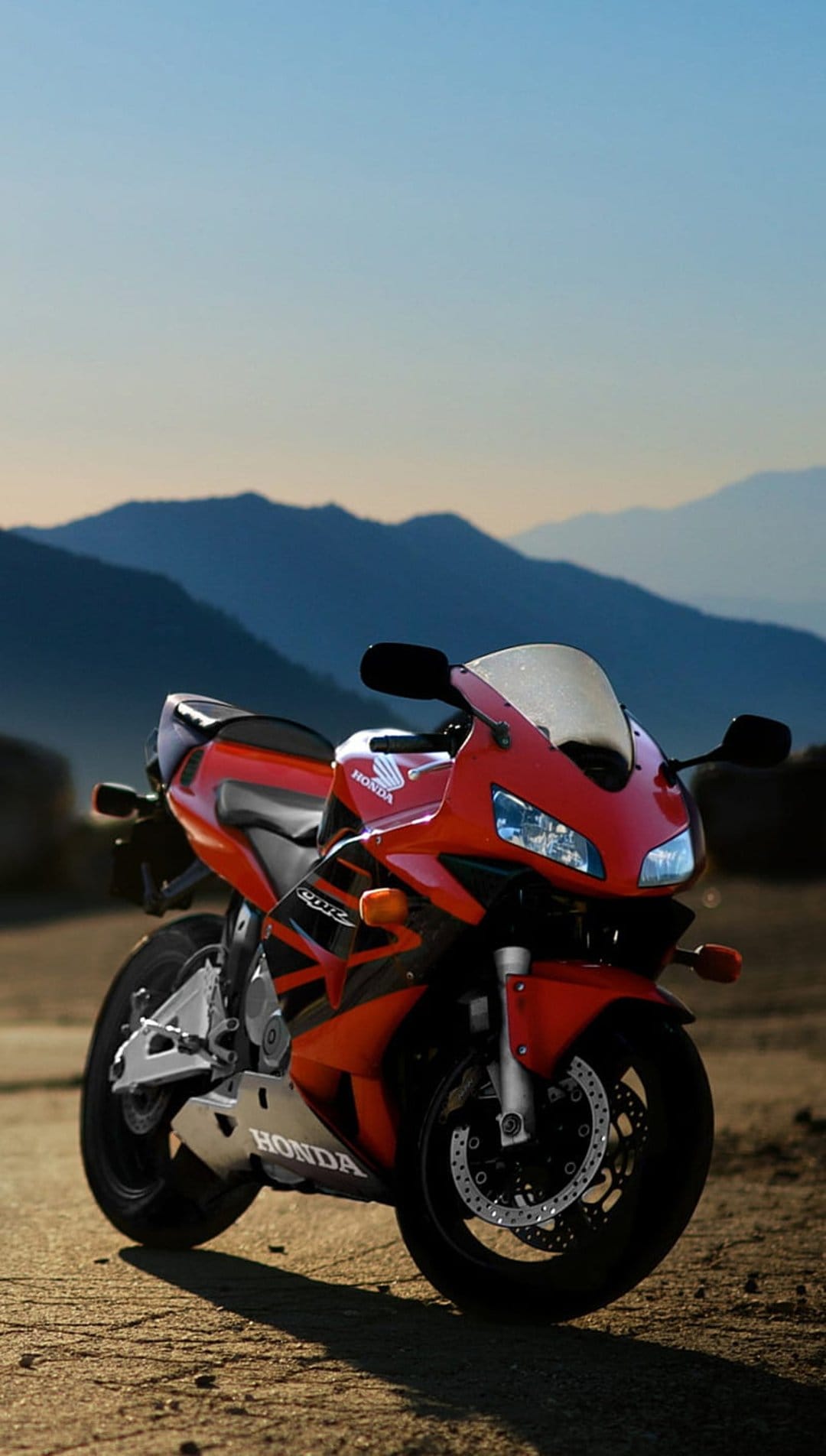 Red Honda CBR motorcycle parked on a rocky mountain ridge with a blue sky background in vertical orientation.
