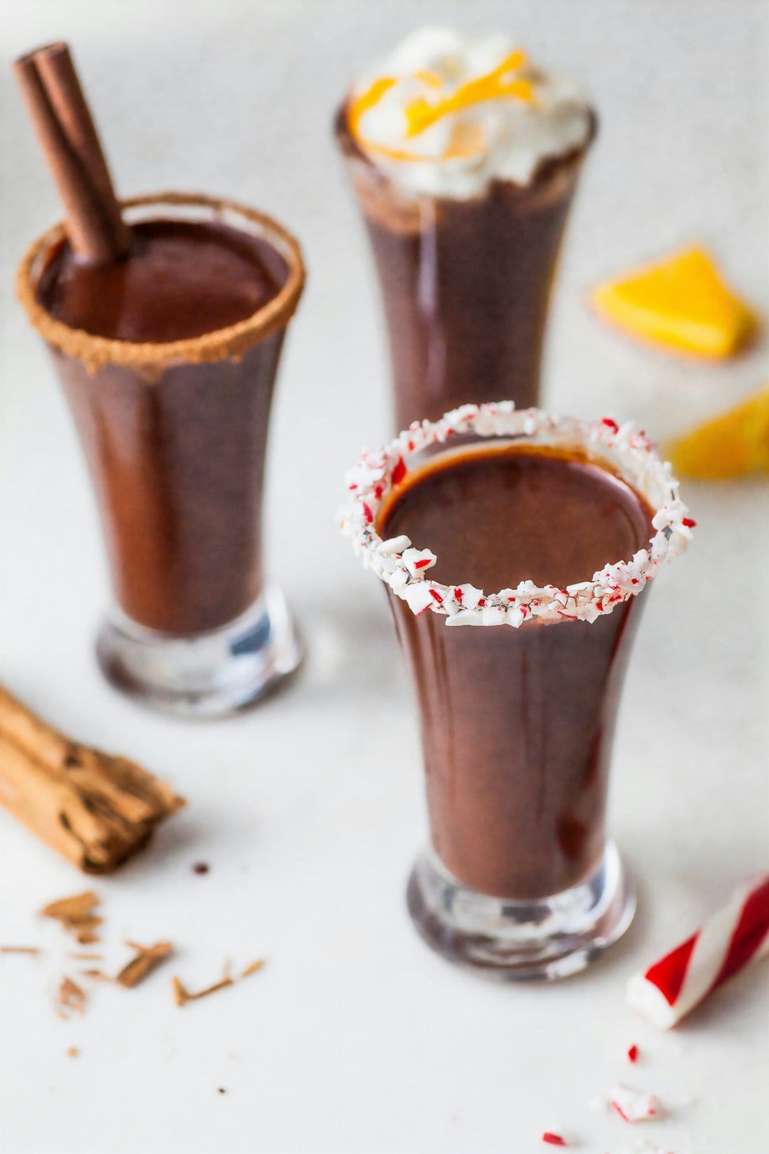 Three cocoa shot glasses with cinnamon sticks, peppermint, and orange zest against a festive background.