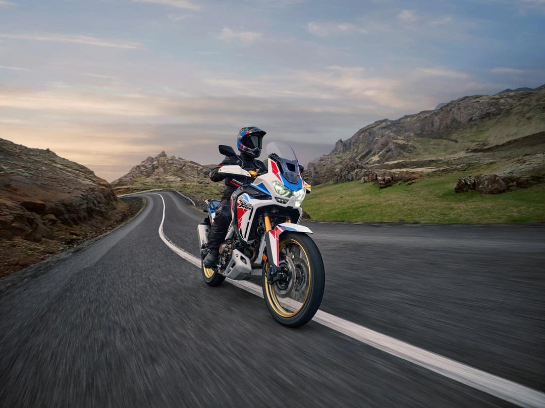 White and blue adventure motorcycle rider leans into a curve on a winding mountain road under a soft dusk sky.