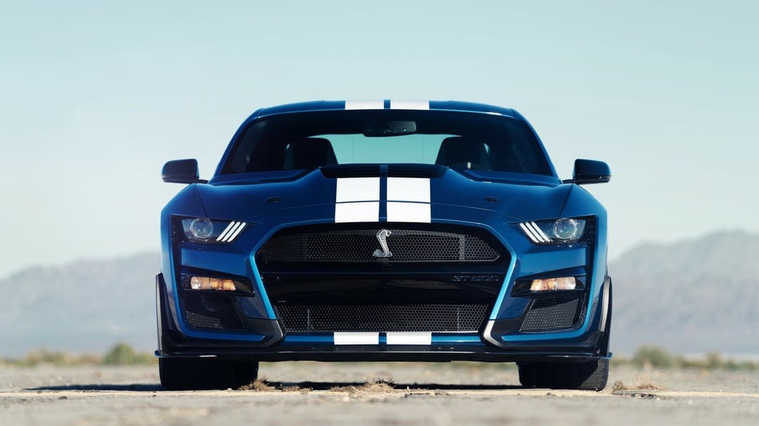 Blue Shelby GT500 muscle car with white racing stripes and glowing headlights against a desert background.