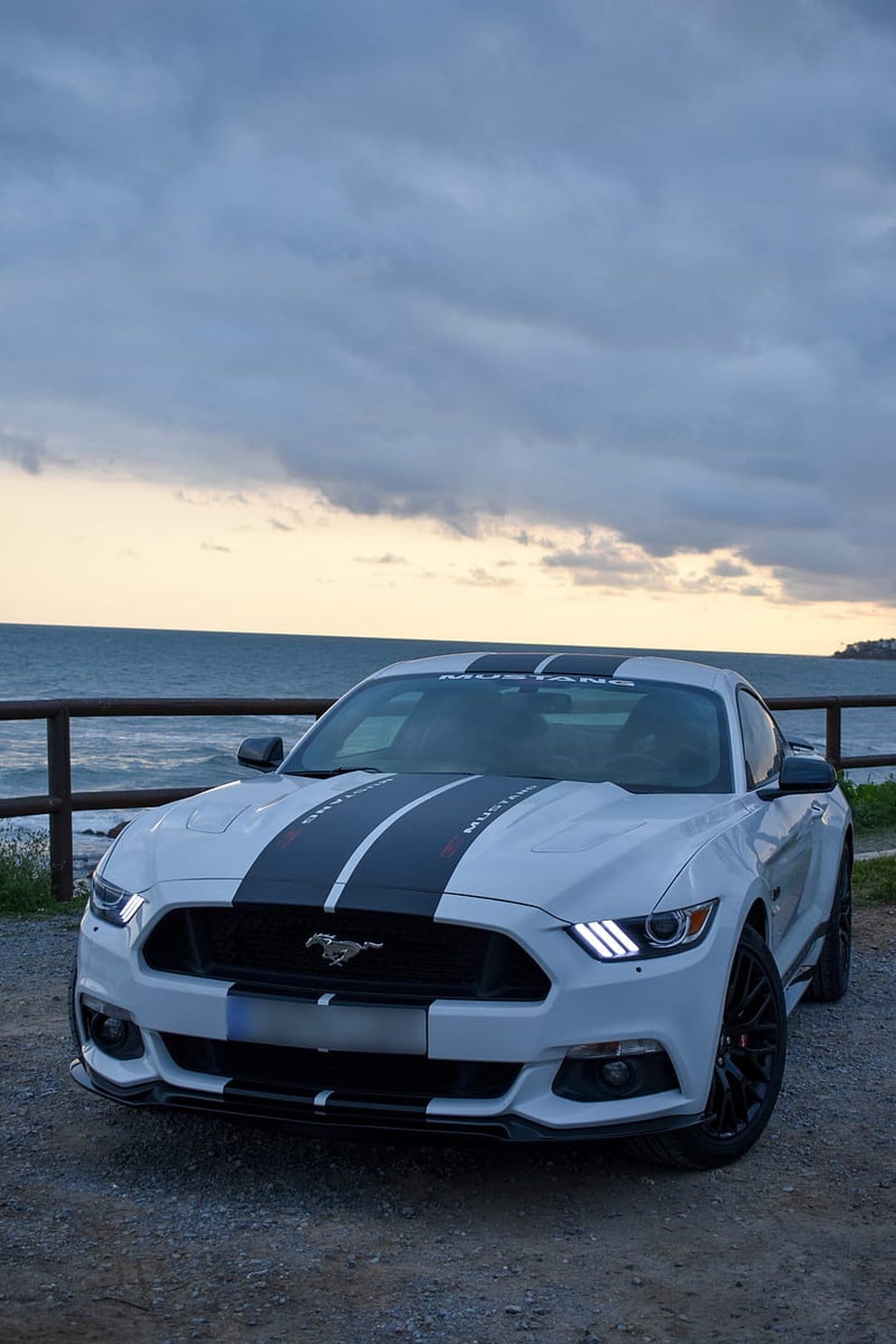 White Ford Mustang with black racing stripes and lit headlights parked on gravel by a stormy sunset ocean view.