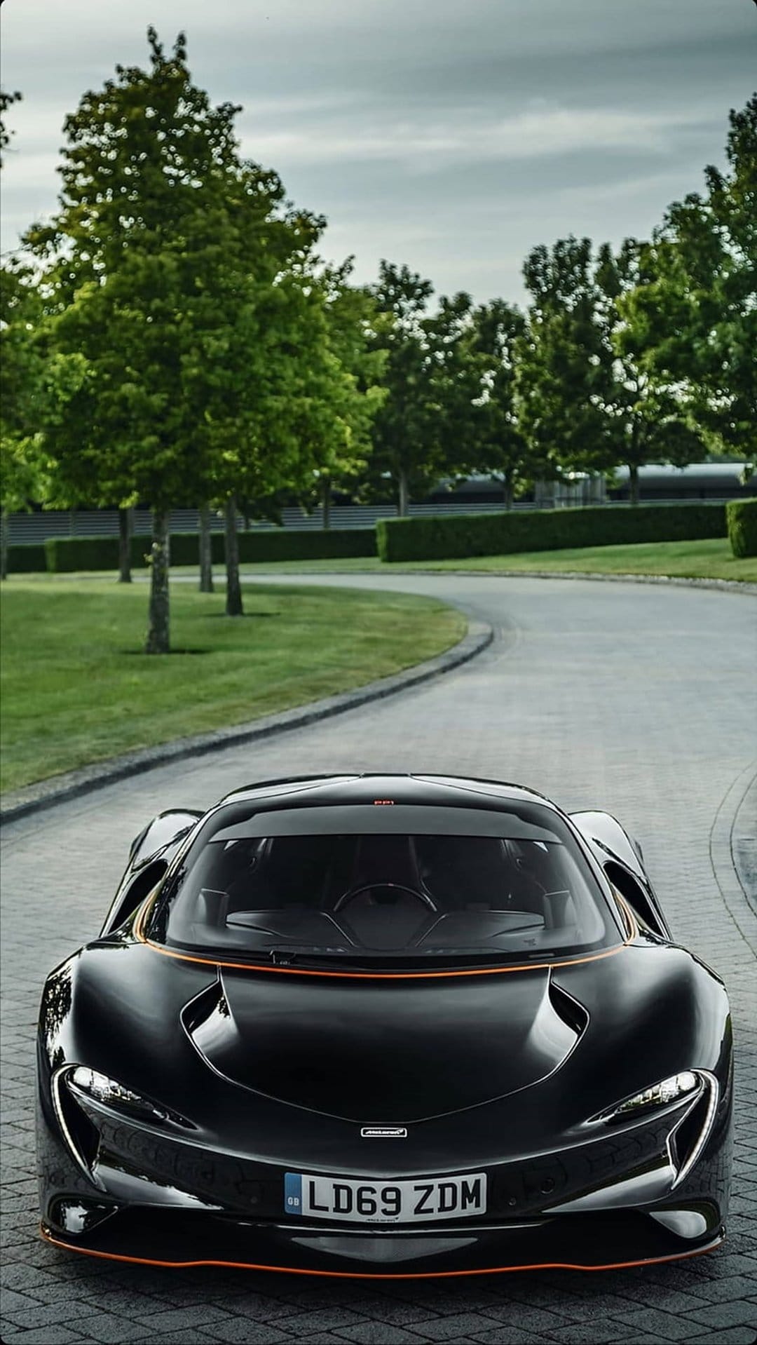 Black McLaren P1 supercar with orange accents on a stone driveway surrounded by green hedges and trees.