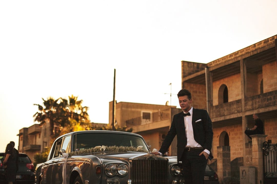 Groom in a black suit leaning against a flower-decorated vintage car near traditional buildings at sunset.