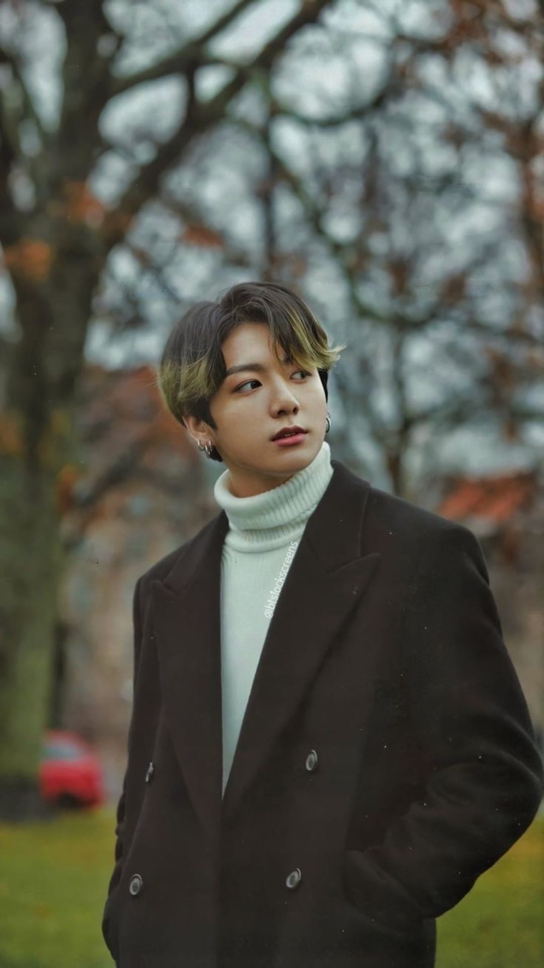 Jeon Jungkook wears a dark coat and blue turtleneck against a blurred autumn background with blonde highlights.