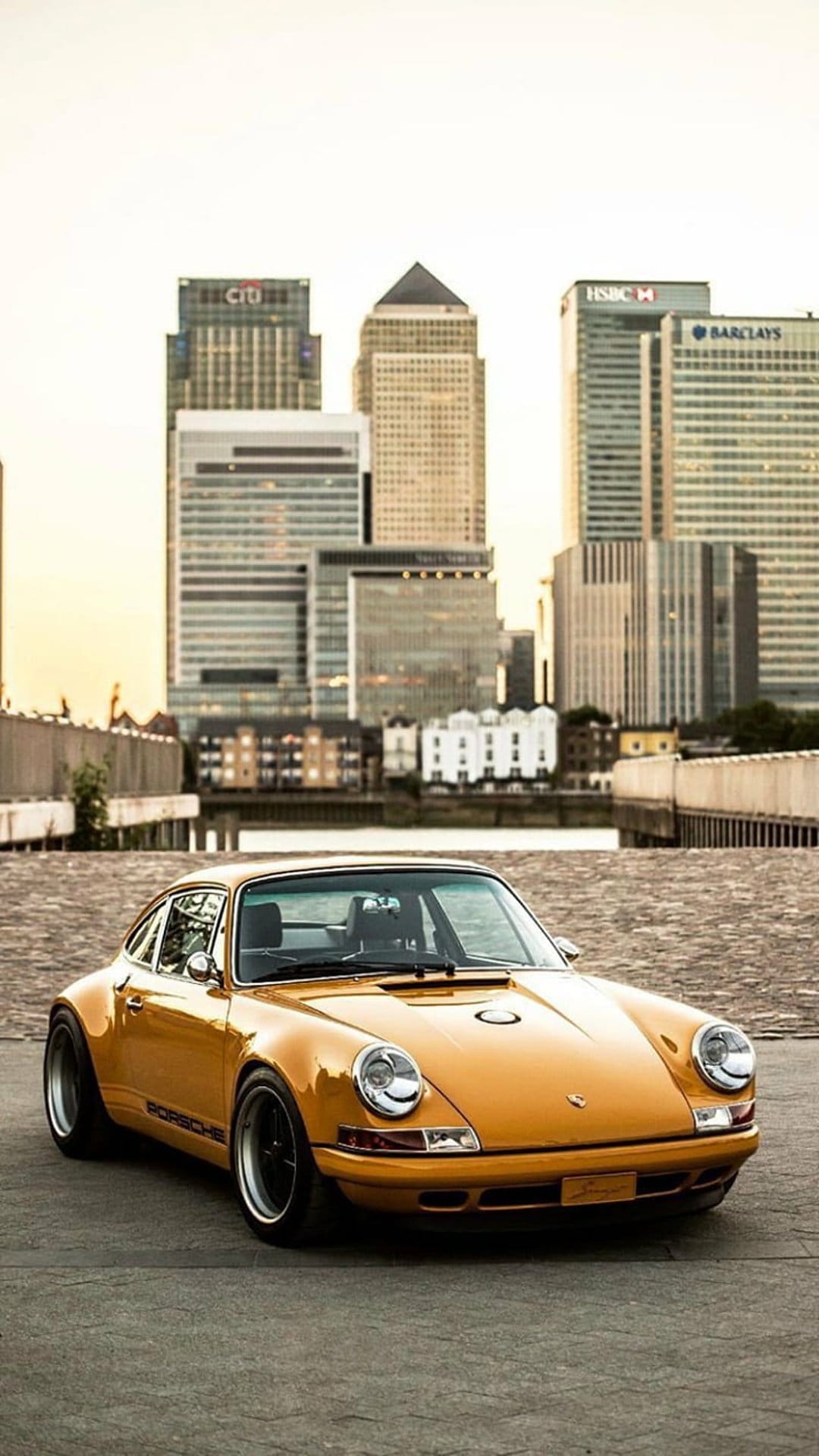 Orange vintage Porsche 911 parked on a paved street with the modern glass towers of Canary Wharf in the background.