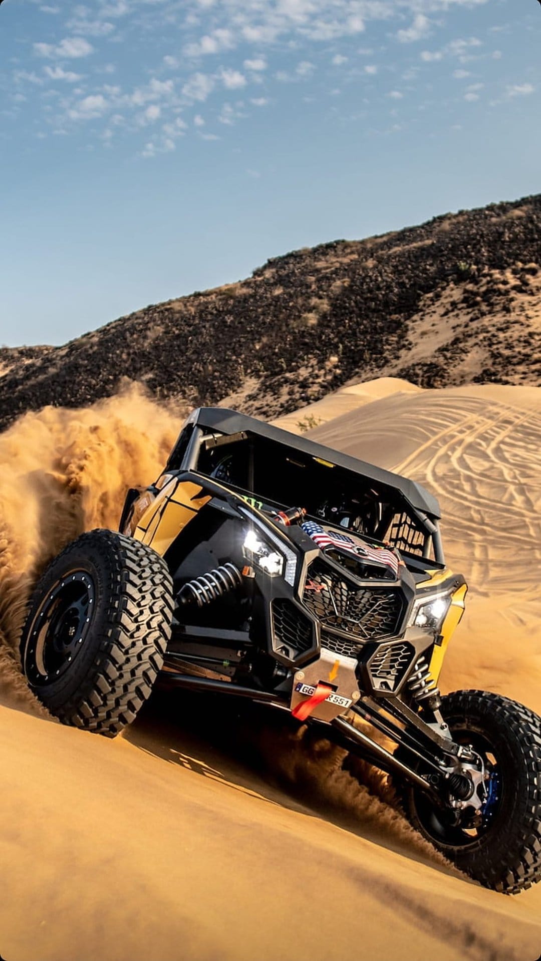 Can-Am Maverick X3 UTV kicking up sand while racing across desert dunes under a bright blue sky.