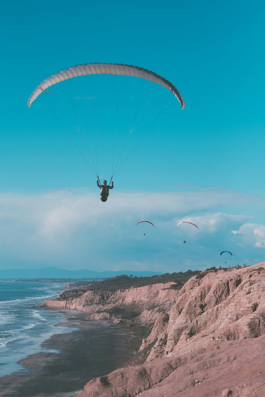 Paragliders float in a blue sky above steep rocky cliffs and ocean waves crashing on a sandy shoreline below.