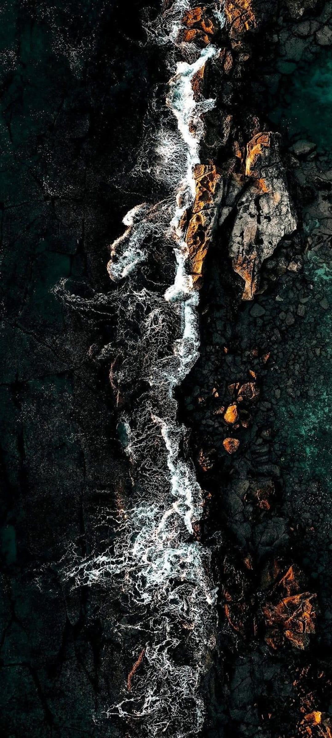 White waves hit jagged black volcanic rocks and emerald sea water seen from a top-down aerial perspective.