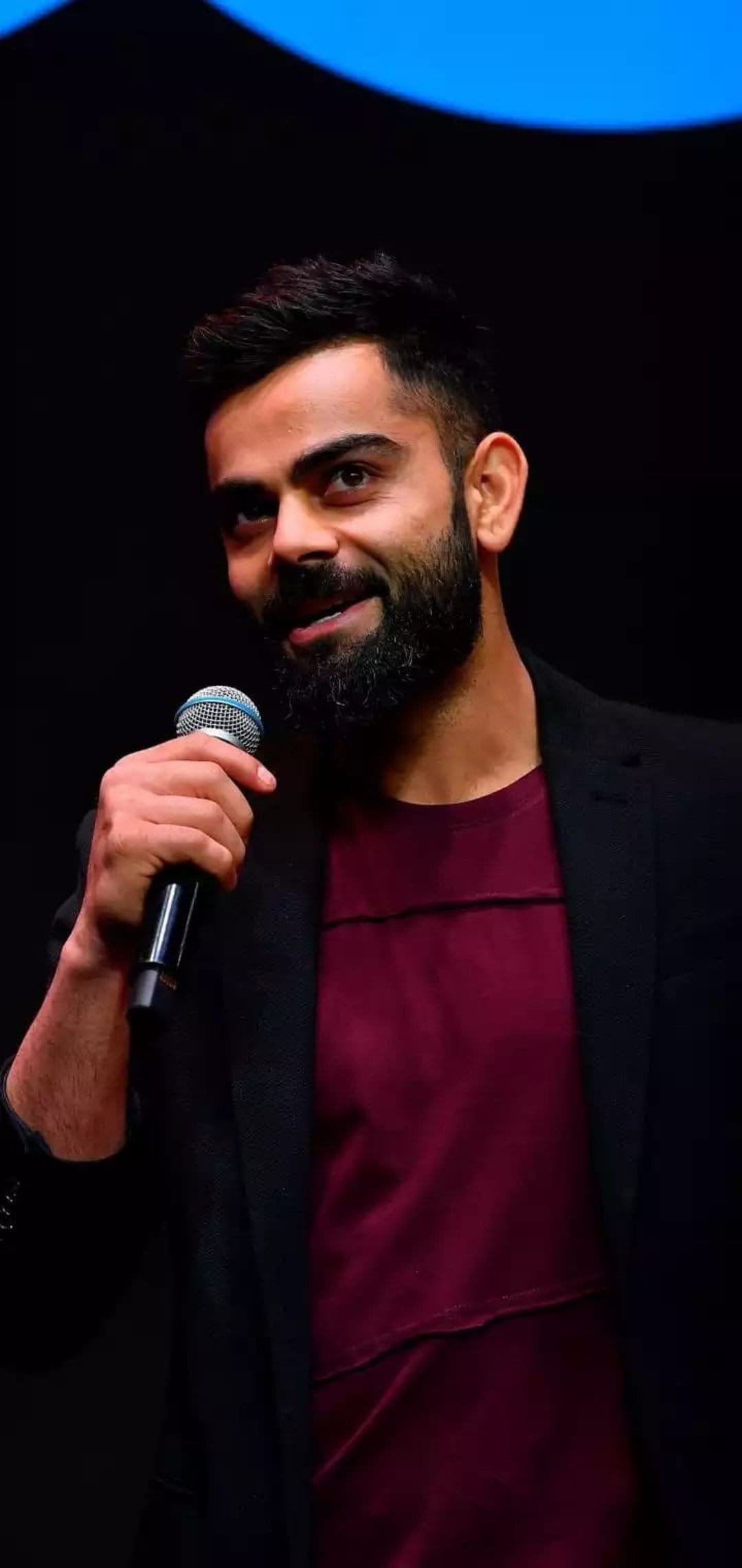 Virat Kohli holds a microphone wearing a dark blazer and maroon t-shirt against a minimalist dark background.