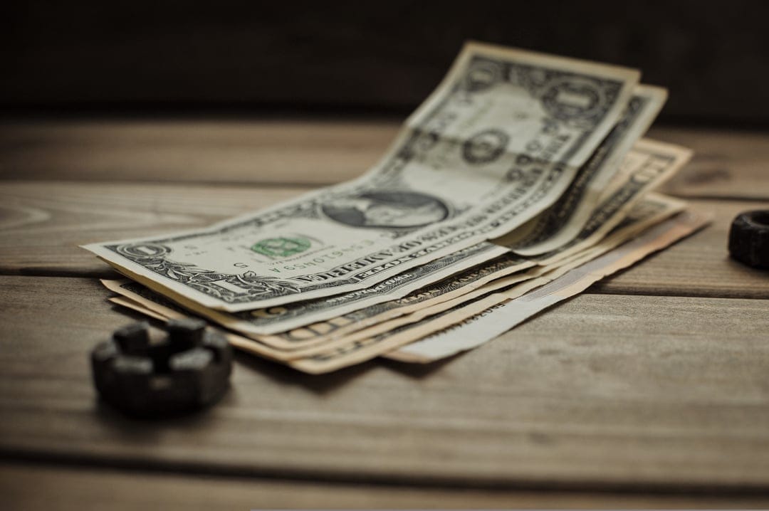 Fanned US dollar bills and a small metallic nut sit on a textured rustic wooden table under soft warm lighting.