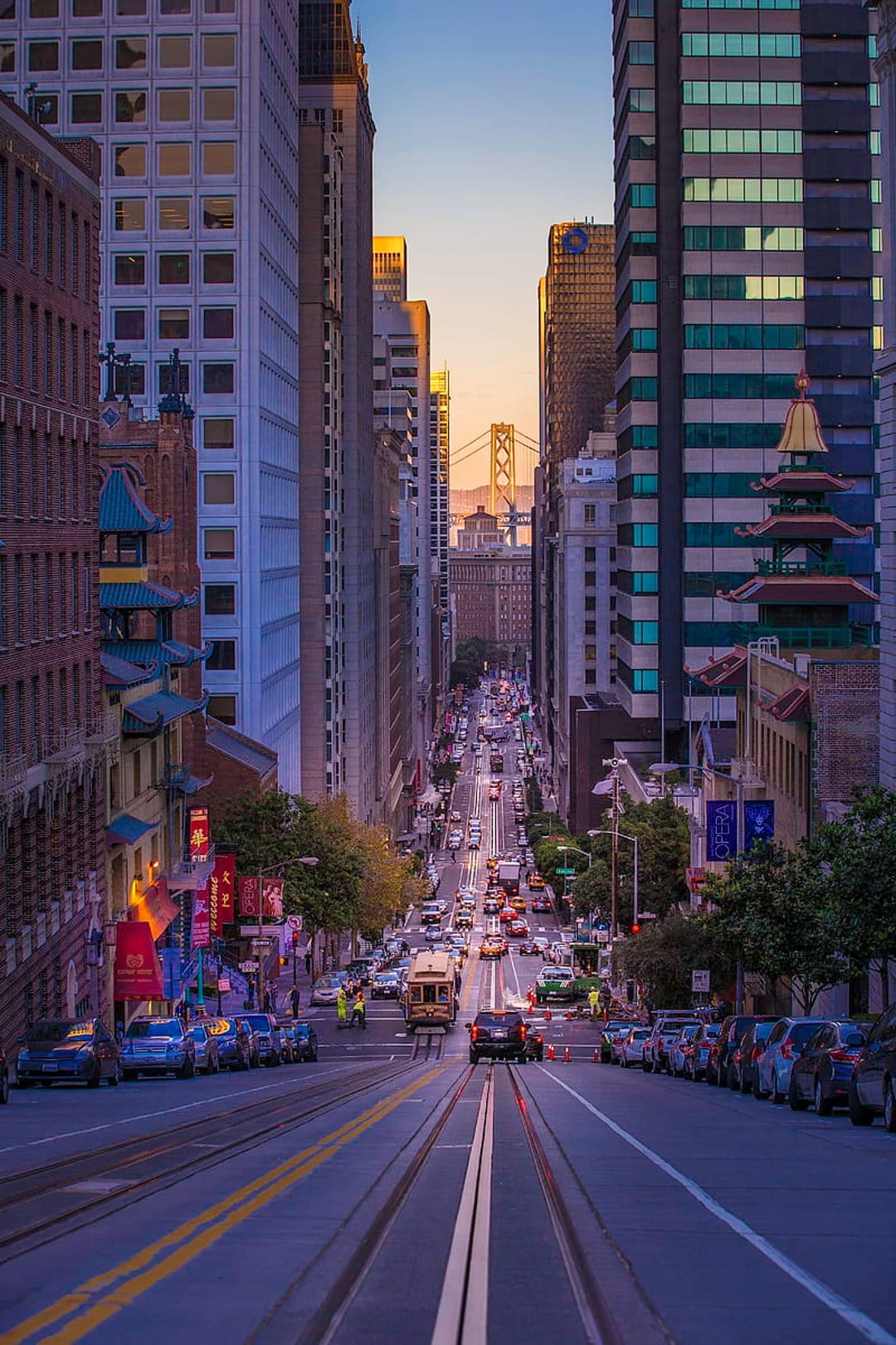 Cable car descending a steep city hill during a bright orange sunset with towering buildings on both sides.