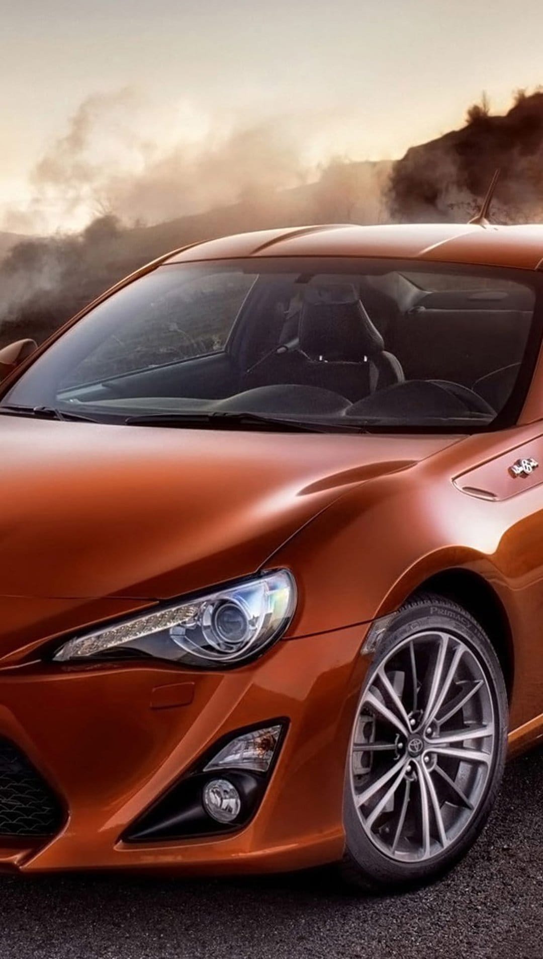 Orange Toyota GT86 sports car with glowing headlights on a mountain road during a misty sunset at golden hour.