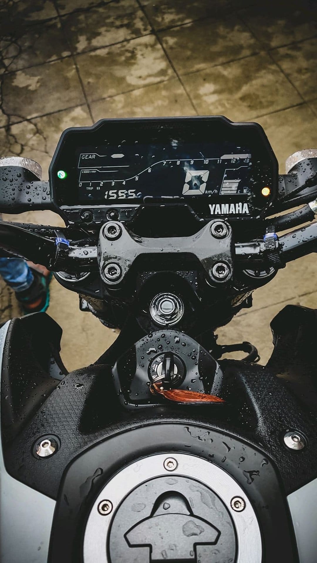 Yamaha motorcycle digital dashboard covered in raindrops with a key in the ignition and chrome fuel cap details.