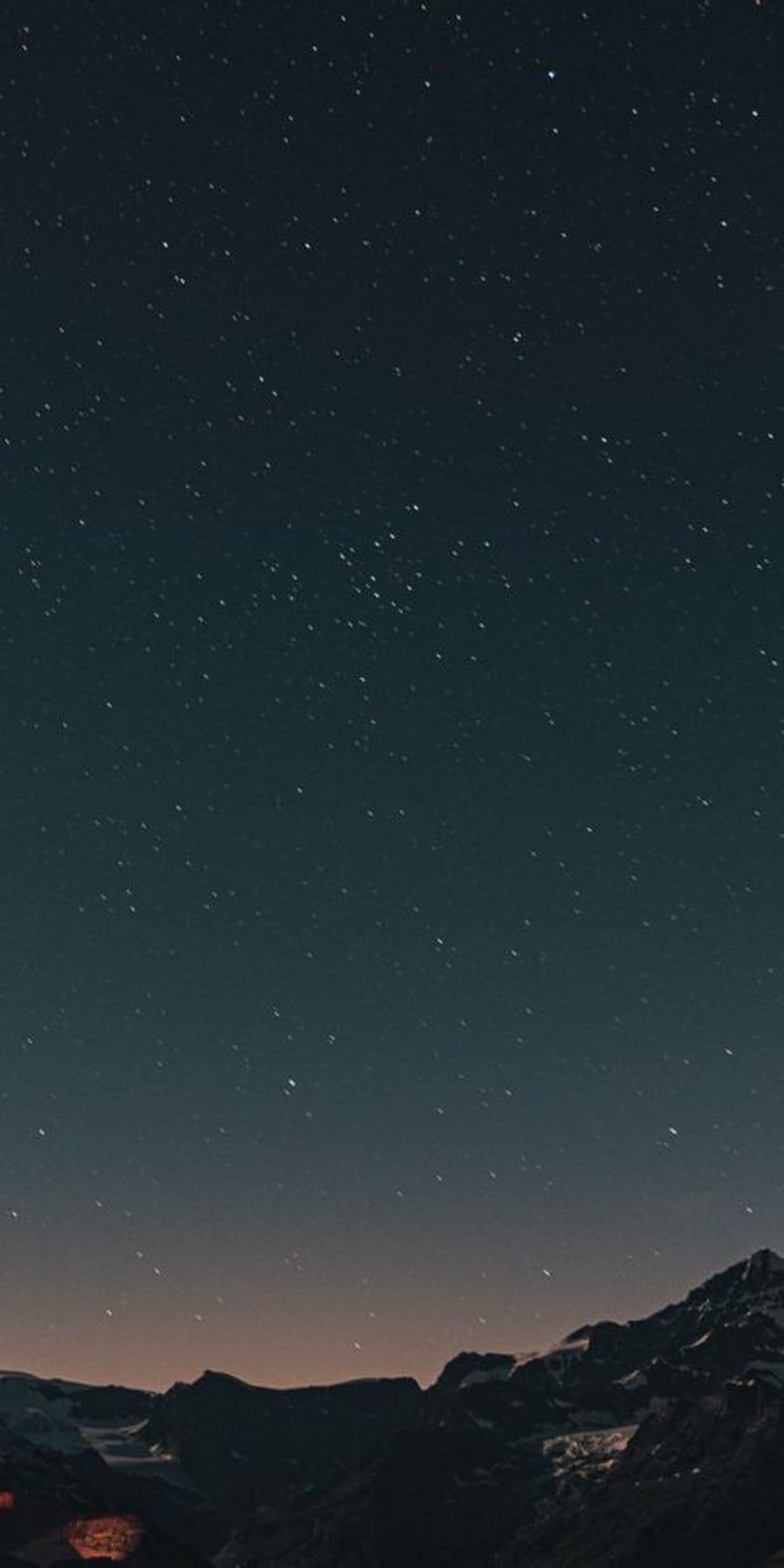 Dark mountain peaks silhouetted against a dense field of stars with a soft light glow on the distant horizon.