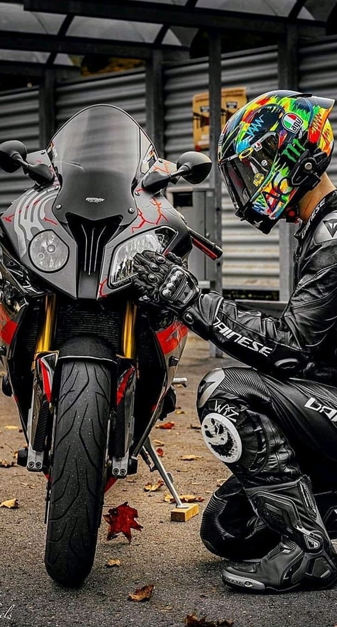 Motorcyclist in a racing suit touches a red sportbike on a ground covered with fallen autumn leaves.