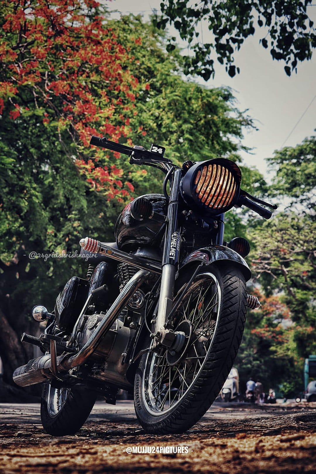 Vintage black motorcycle with a ribbed headlight parked near vibrant red foliage and green trees at a low angle.