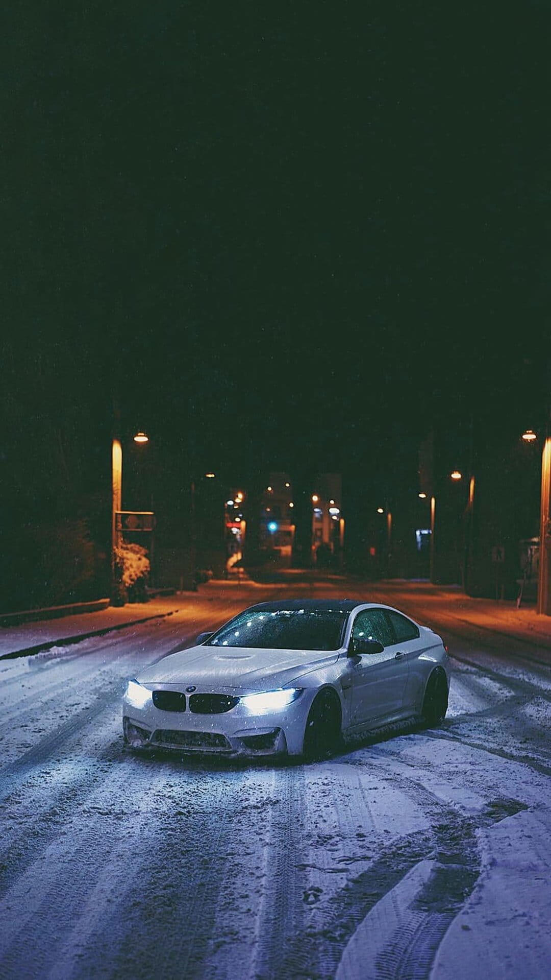 BMW M4 drifts on a snowy street at night with glowing headlights and orange streetlights in the background.