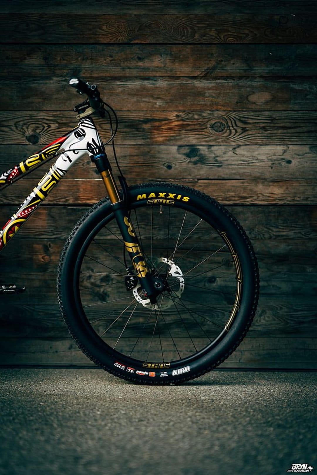 White mountain bike with red tribal graphics and gold spokes leaning on a rustic wooden wall in vertical view.