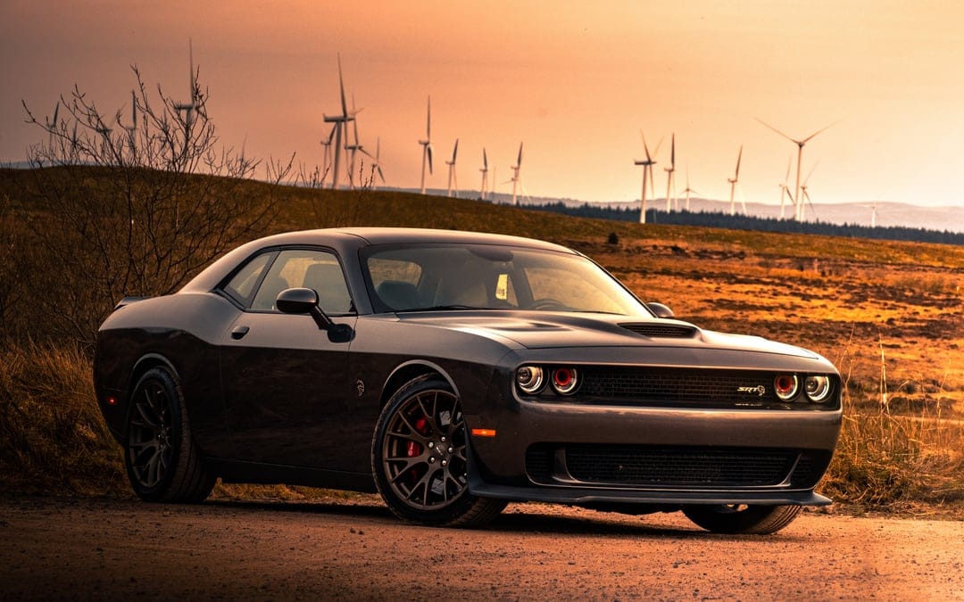 Dark grey muscle car parked on a dirt path against a golden sunset and wind turbines on a distant hill.