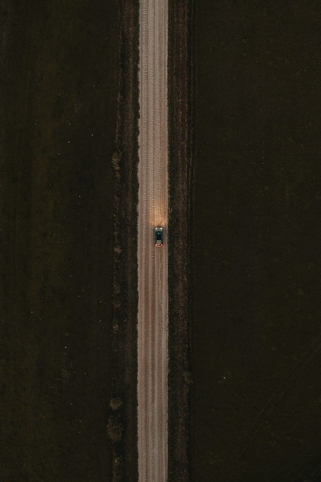 White headlights of a car illuminate a dusty brown road from an overhead drone perspective in a dark landscape.