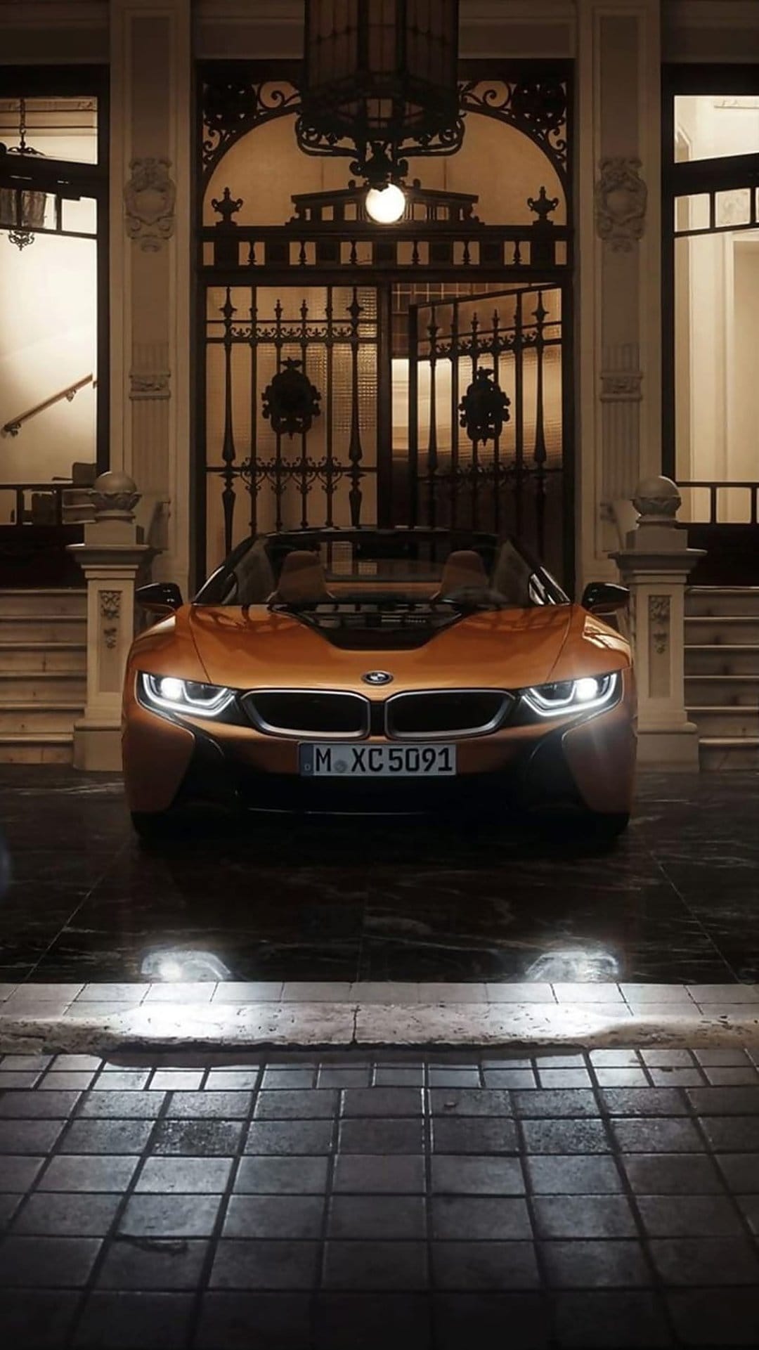 Orange BMW i8 Roadster parked on wet pavement before ornate iron gates and stone stairs with glowing headlights.