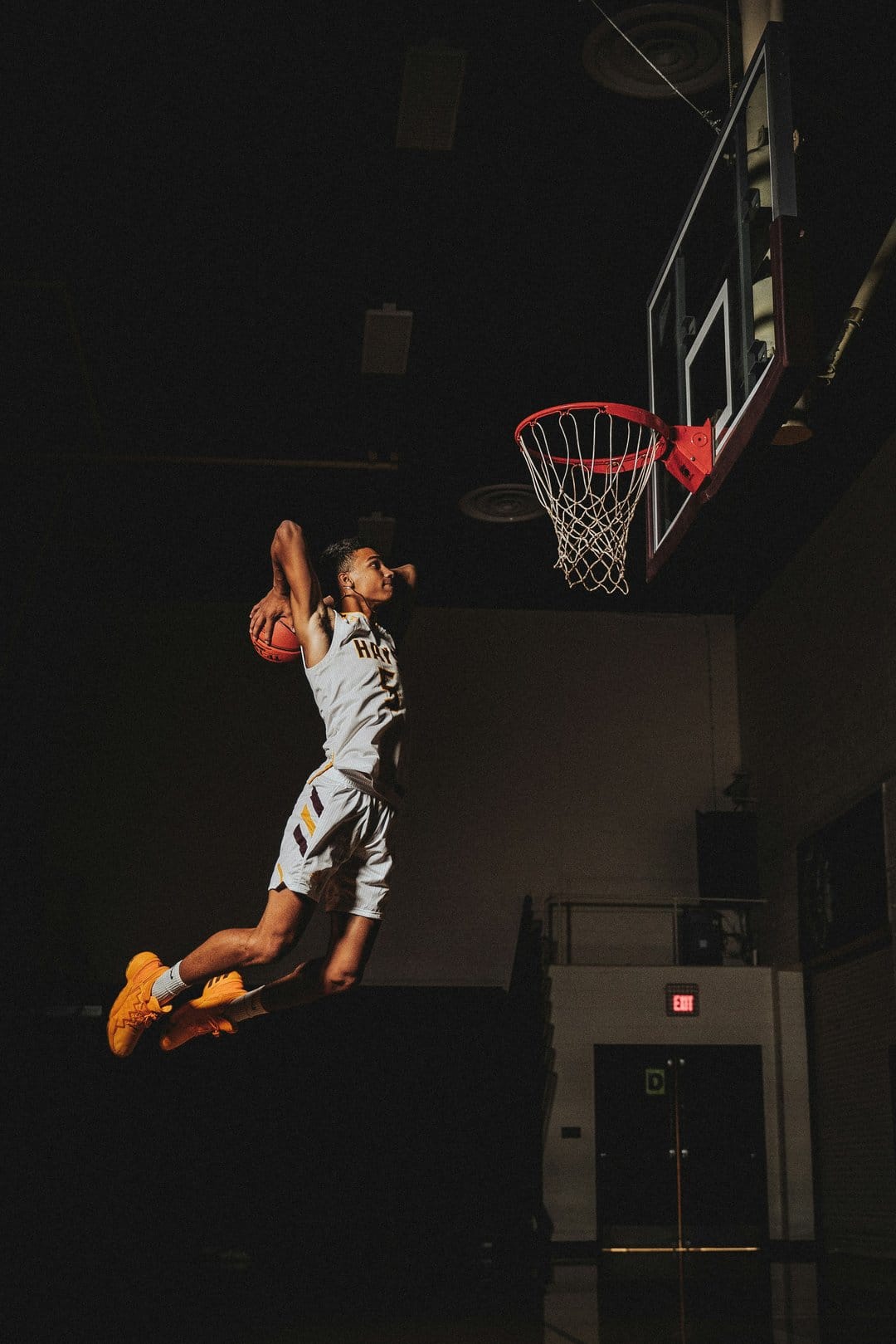 Basketball player in a white and yellow jersey mid-dunk against a dark gym background with dramatic lighting.