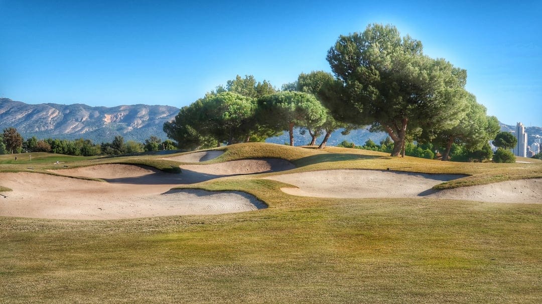 Sculpted golf bunkers and lush fairways lead toward distant mountains and a city skyline under a clear blue sky.