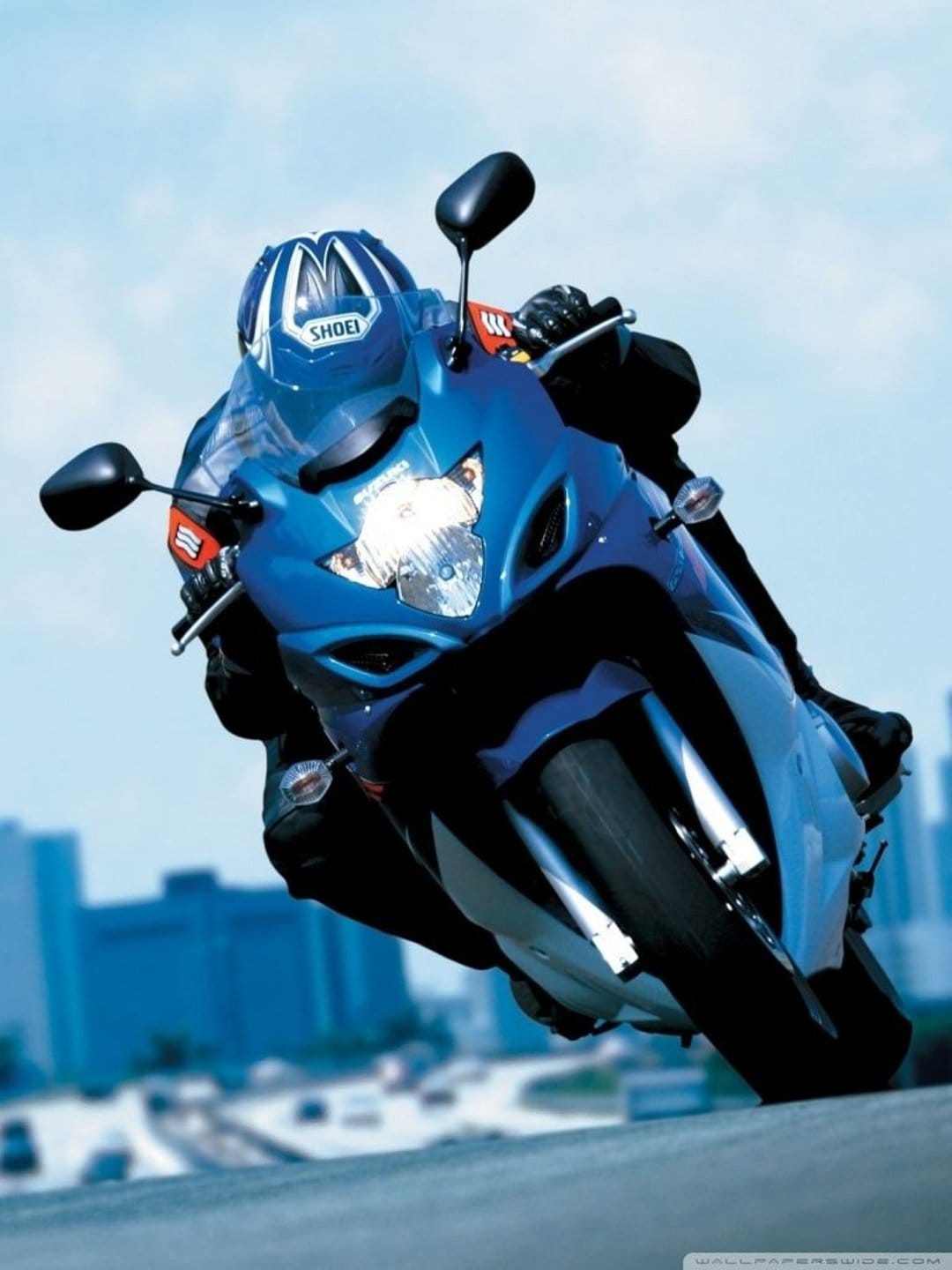 Blue sport motorcycle rider in a racing suit leans into a turn against a blurred cityscape and sky.