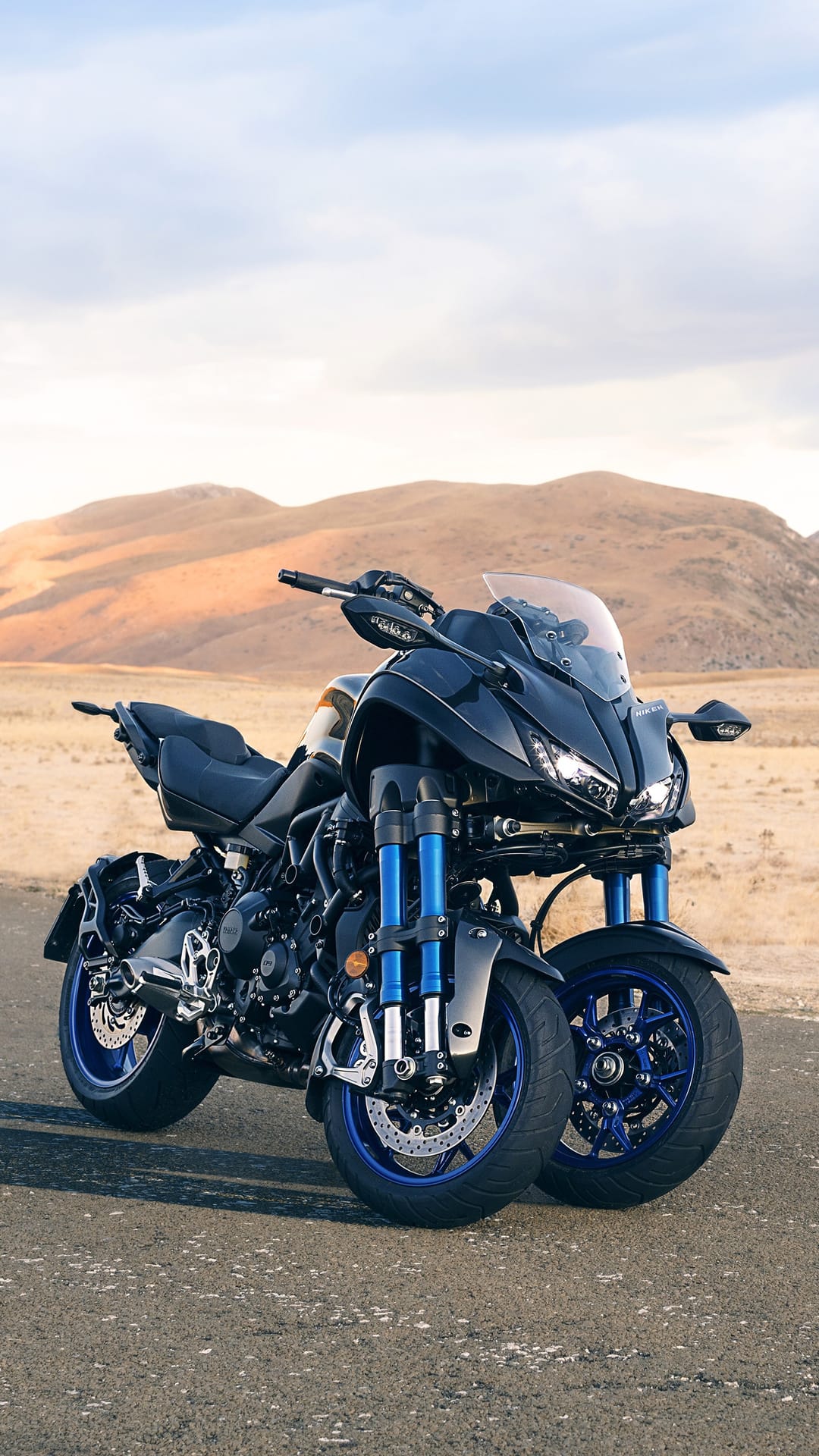 Yamaha Niken GT three-wheeled motorcycle on a desert asphalt road under a cloudy sky with blue suspension forks.