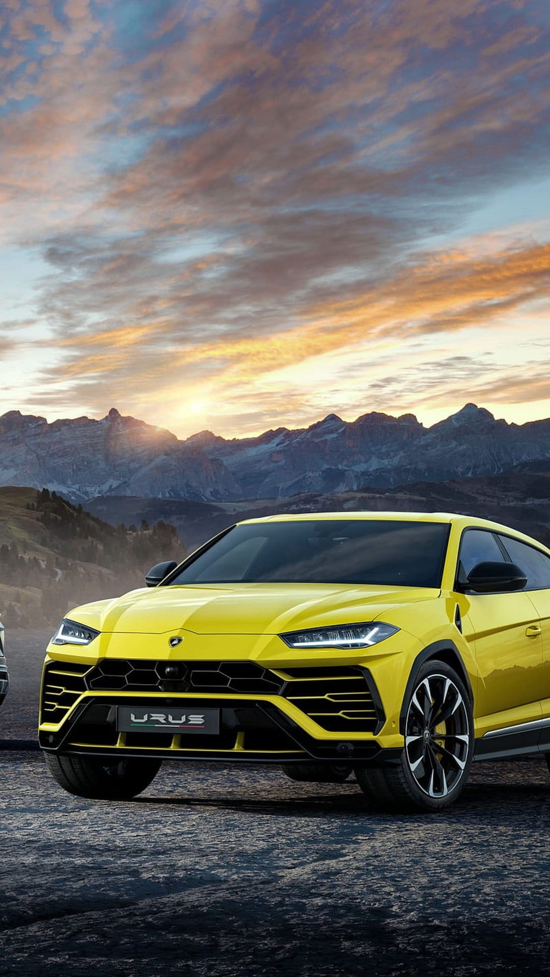Yellow Lamborghini Urus SUV parked on rugged rocks with a colorful sunset and mountain silhouettes in the distance.