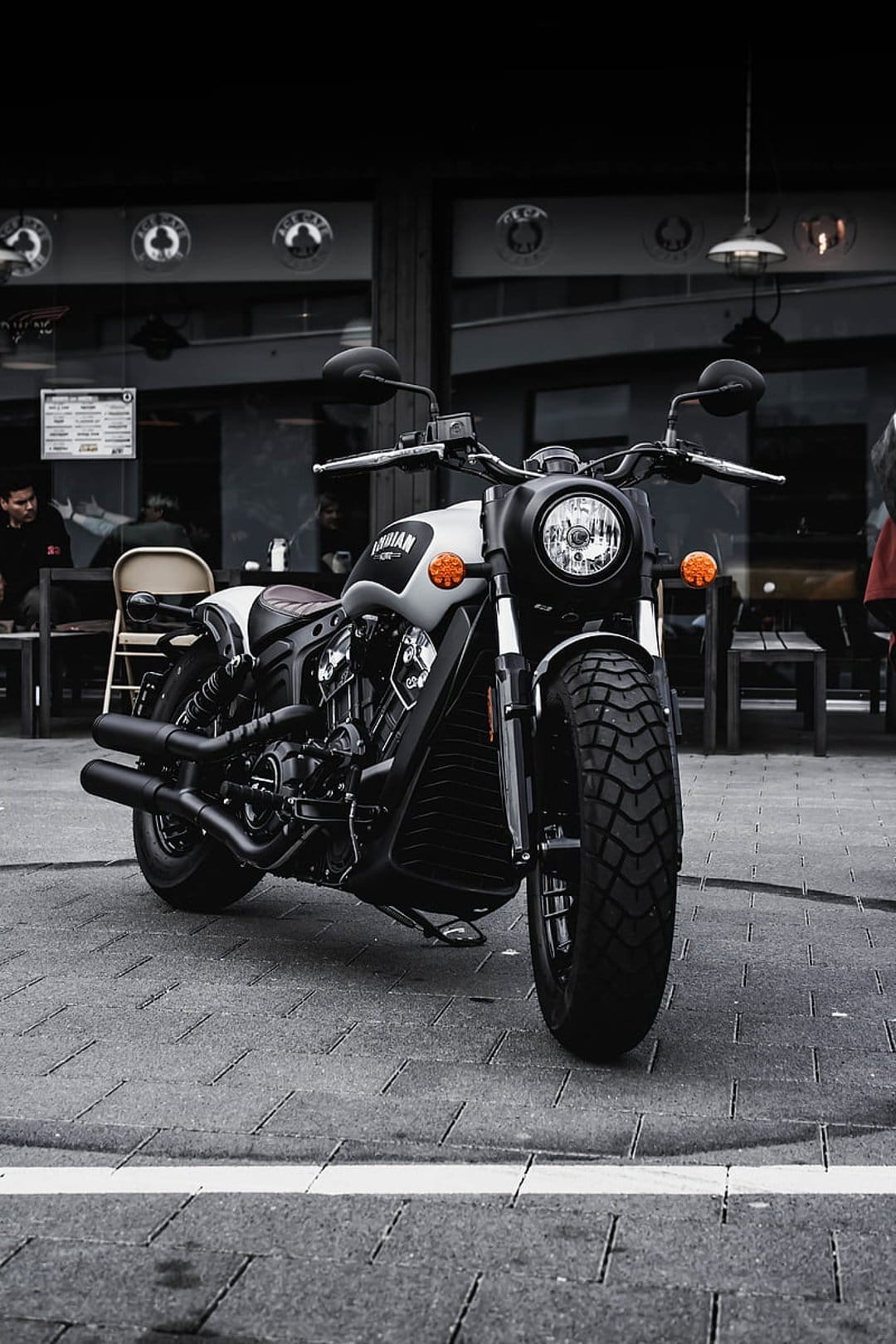 Indian motorcycle with matte grey finish parked on cobblestone street near a blurred cafe with outdoor seating.