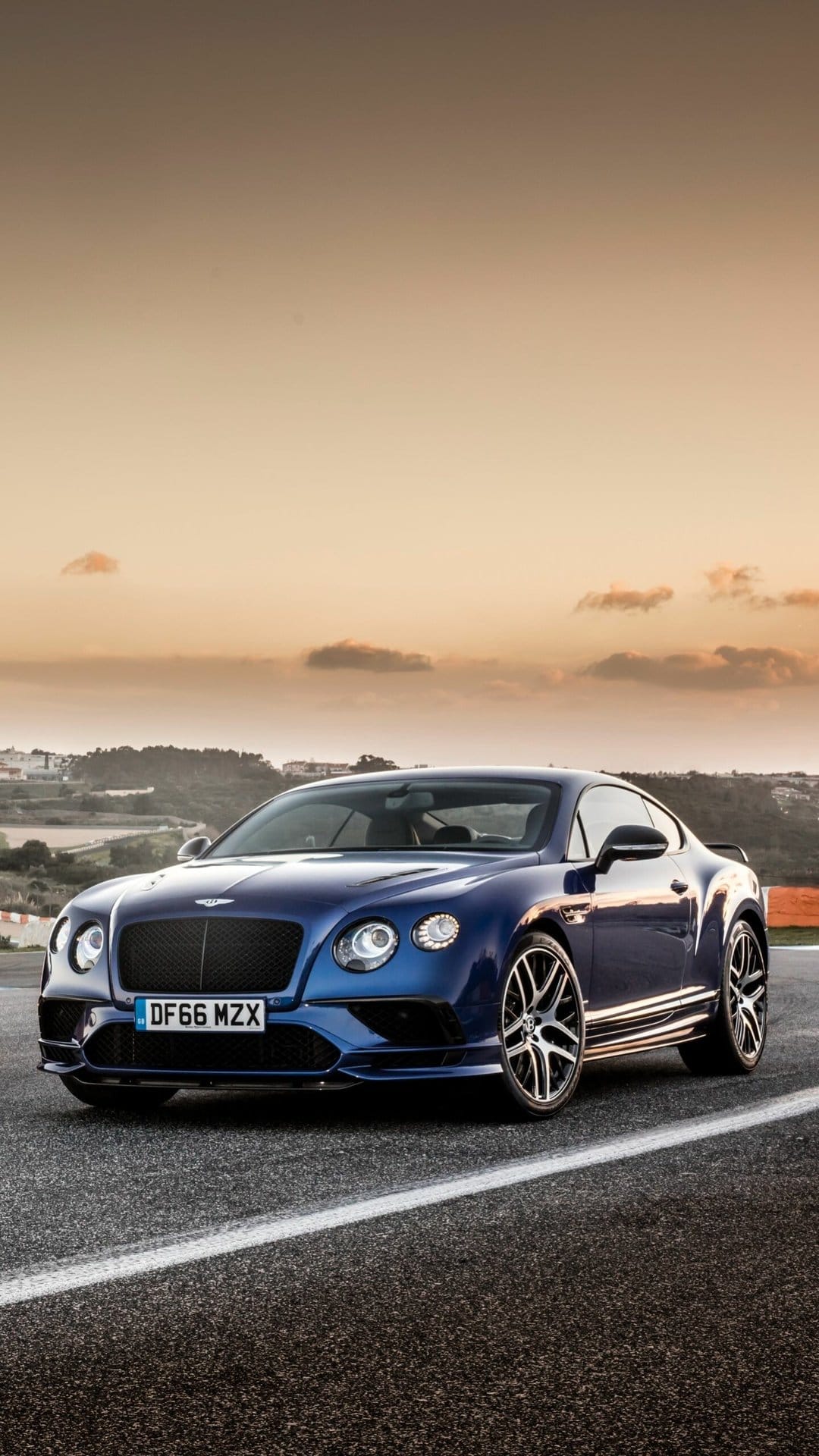 Front view of a dark blue luxury coupe on a paved road under a golden sunset sky with soft clouds.