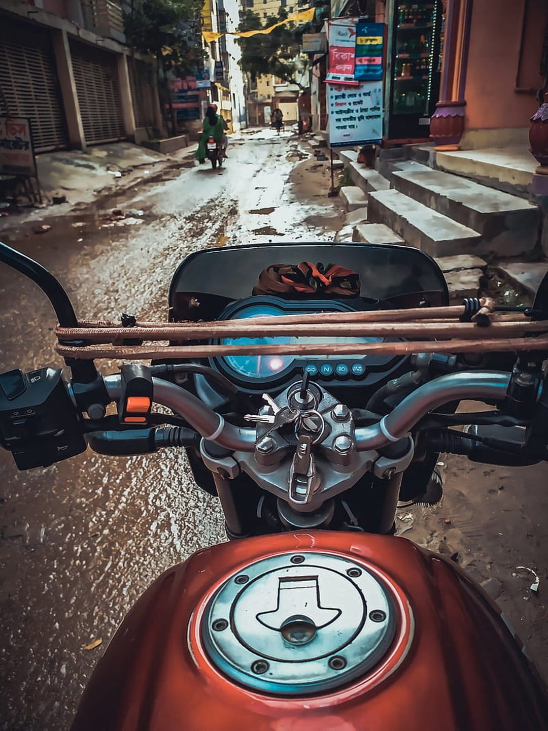 Motorcycle handlebars carrying timber navigate a wet, narrow street lined with buildings during a rainy day.
