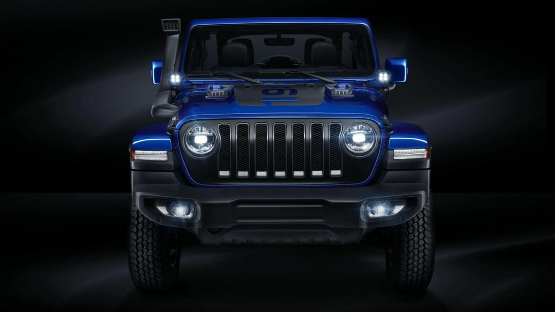 Front profile of a blue Jeep Wrangler with bright LED headlights and off-road gear against a dark background.