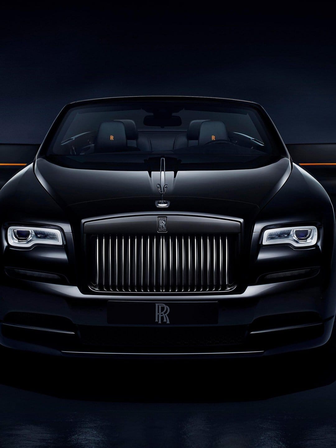 Front view of a black Rolls-Royce with a chrome grille, glowing headlights, and orange headrest embroidery.