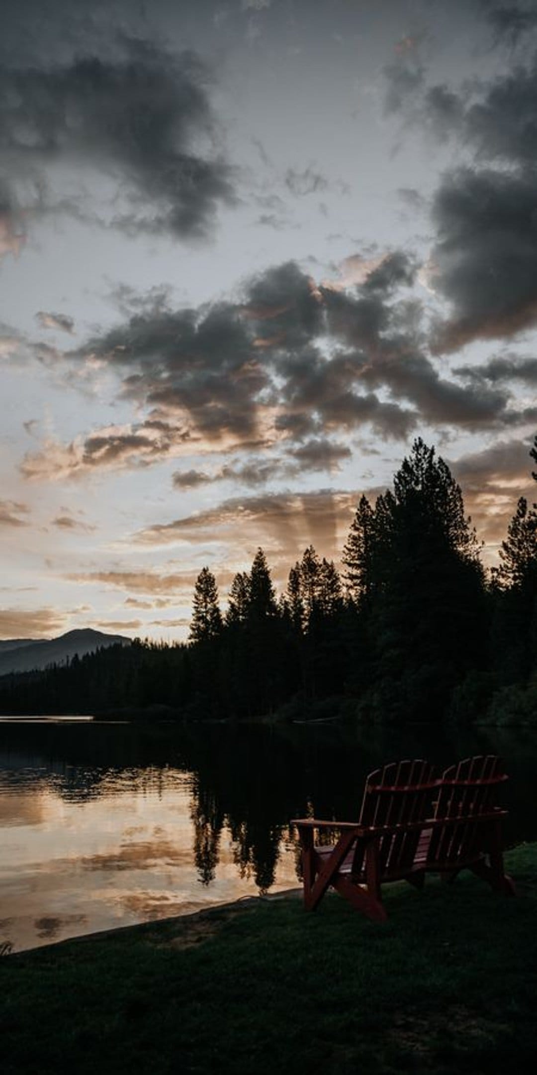 Red Adirondack chairs face a calm lake reflecting a golden sunset with distant mountains and evergreen trees.