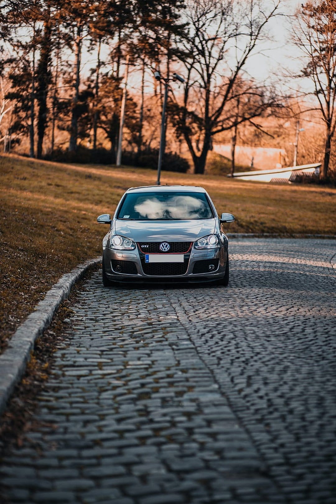 Gray Volkswagen GTI parked on a cobblestone road surrounded by orange autumn trees during a golden hour sunset.