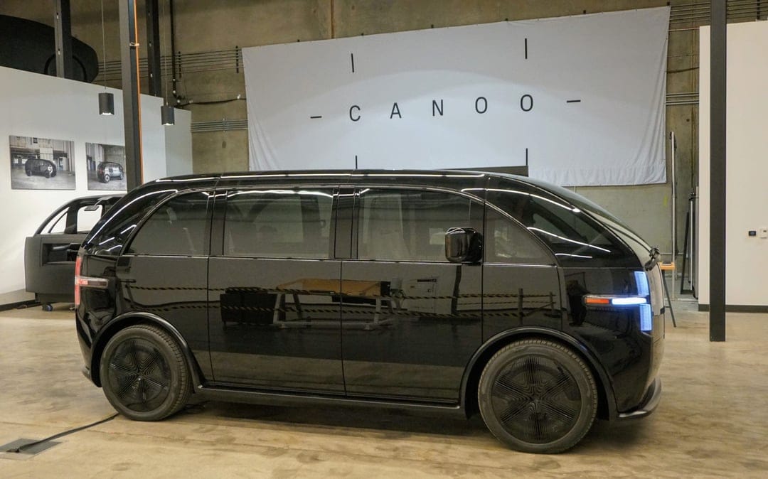 Side profile of a glossy black Canoo electric vehicle in a studio with a white logo banner and concrete floor.