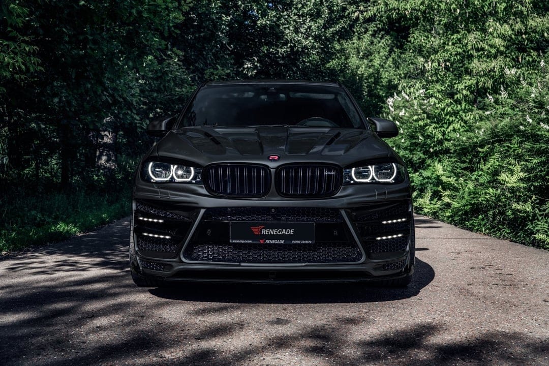 Matte black BMW X5 with Renegade widebody kit and glowing LED headlights positioned on a shadowed forest road.