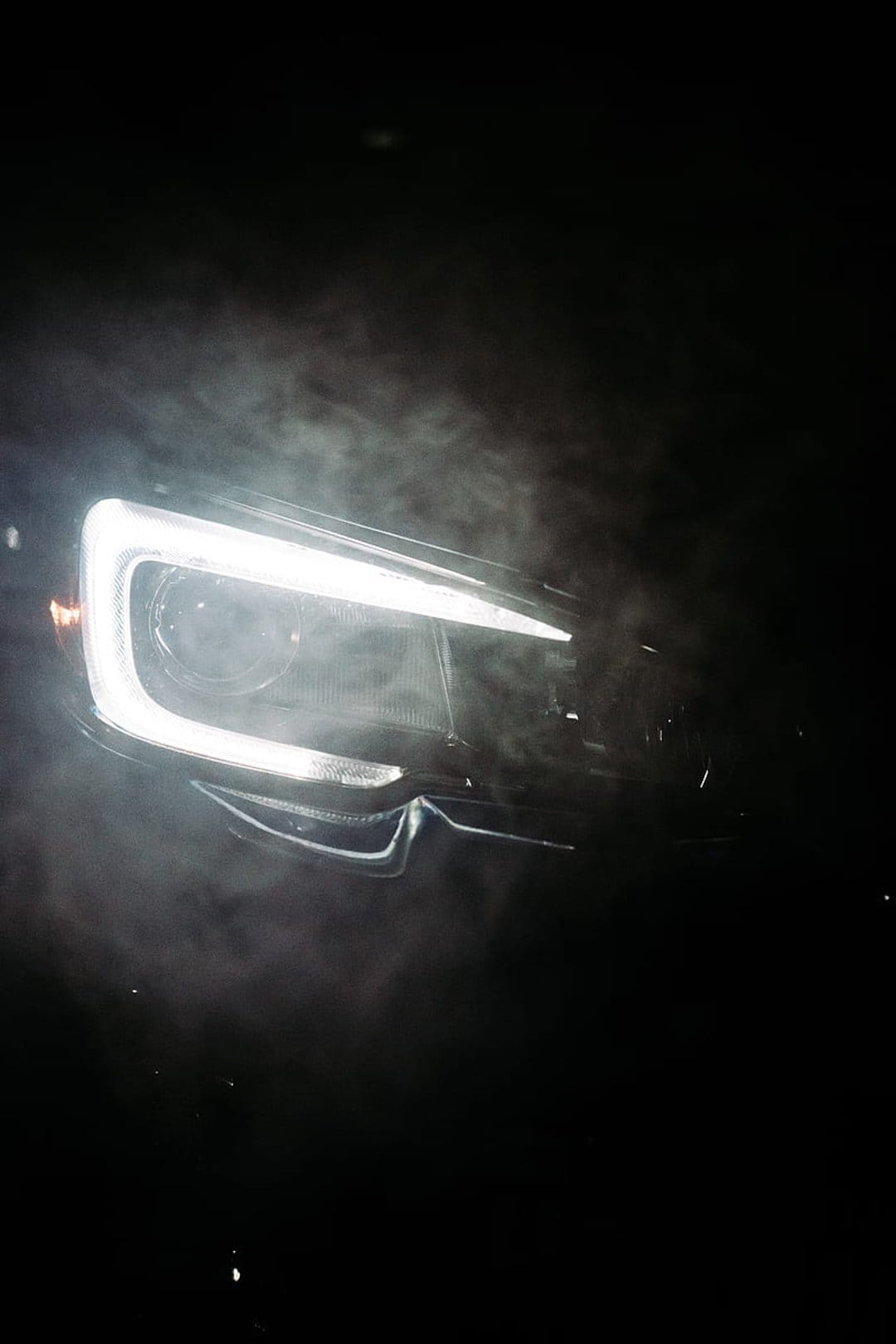 Modern car headlight glowing with bright white light through thick swirling smoke against a black background.