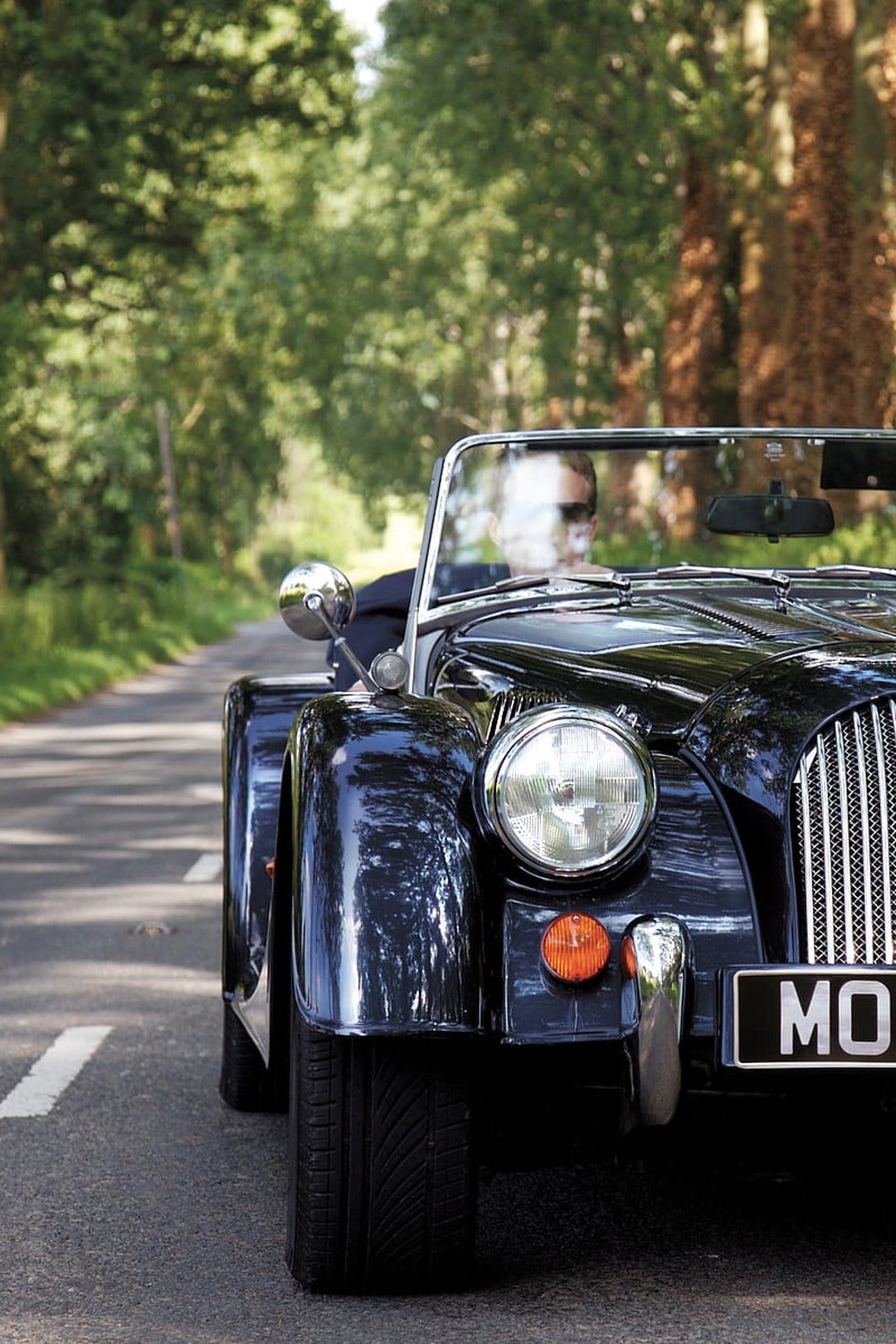 Vintage black convertible car with chrome grille driving through dappled sunlight on a lush tree-lined road.