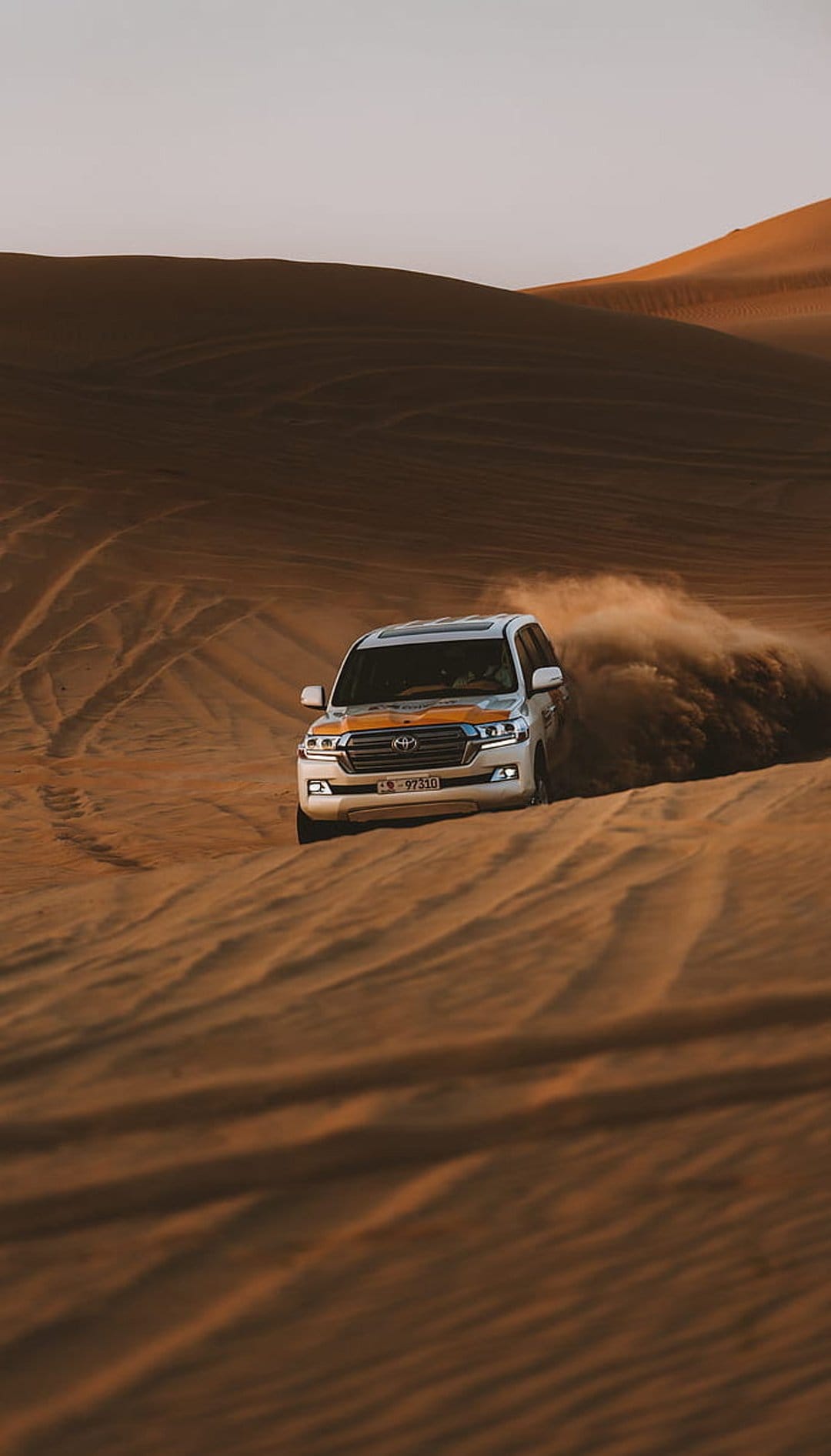 White Toyota Land Cruiser SUV kicking up a large sand cloud while driving across rolling desert dunes.