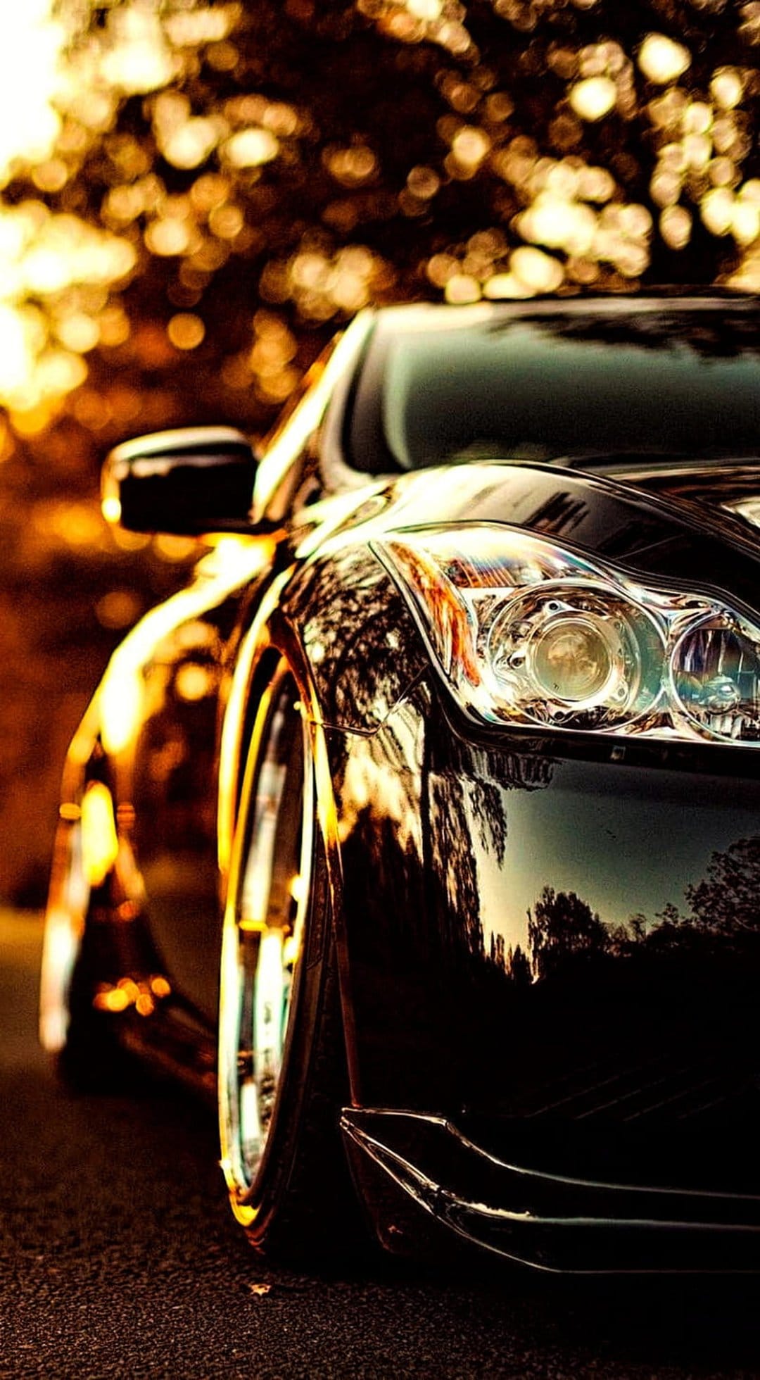 Sleek black sports car headlight reflects golden sunset light and amber bokeh in a close-up detail shot.