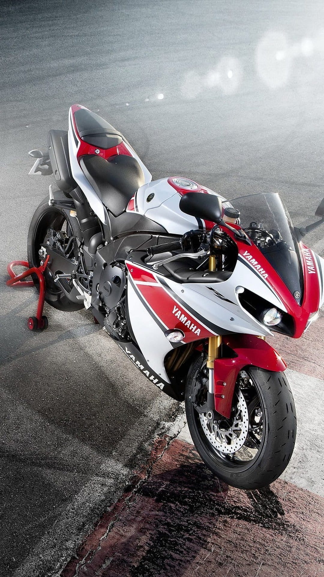 Red and white Yamaha sport motorcycle on a paddock stand atop a grey asphalt racetrack under bright daylight.