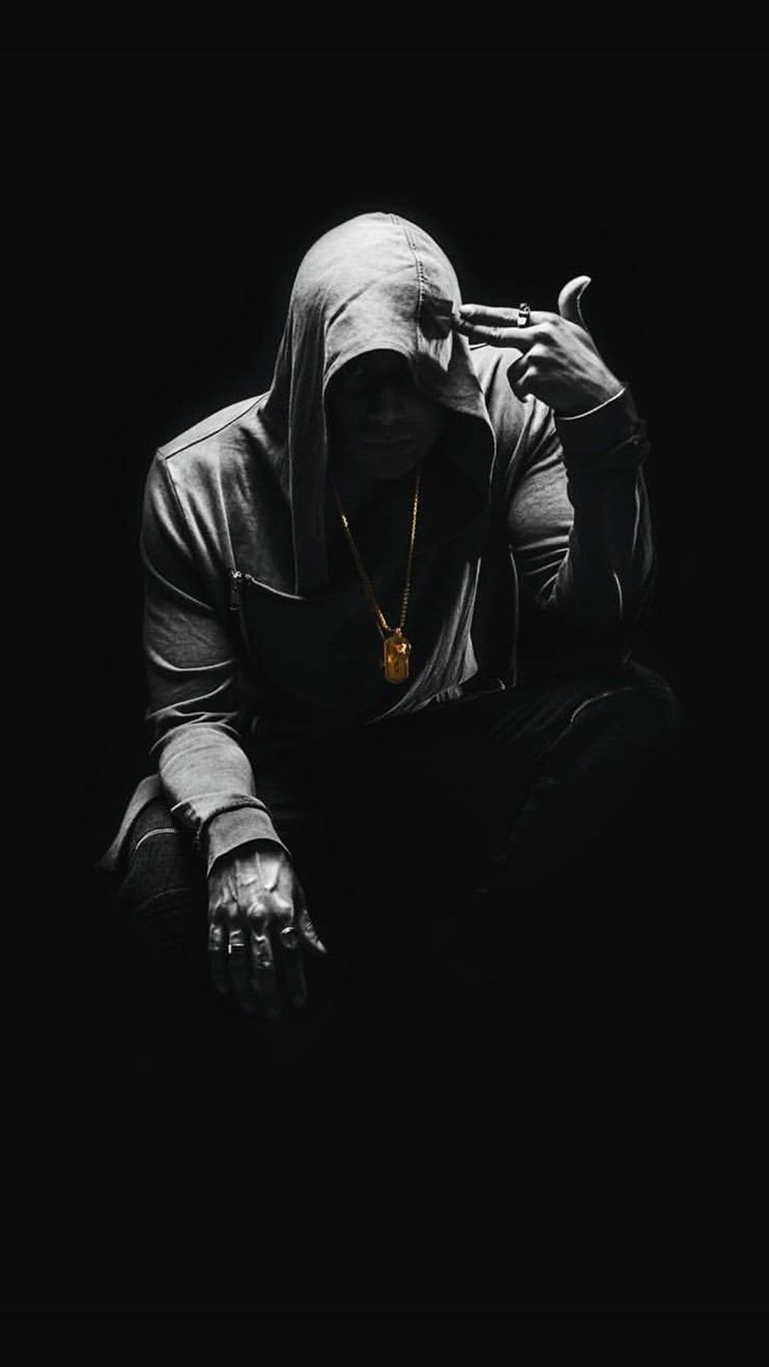 Hooded figure in shadows holds a hand to their temple wearing a gold chain in a monochrome vertical portrait.