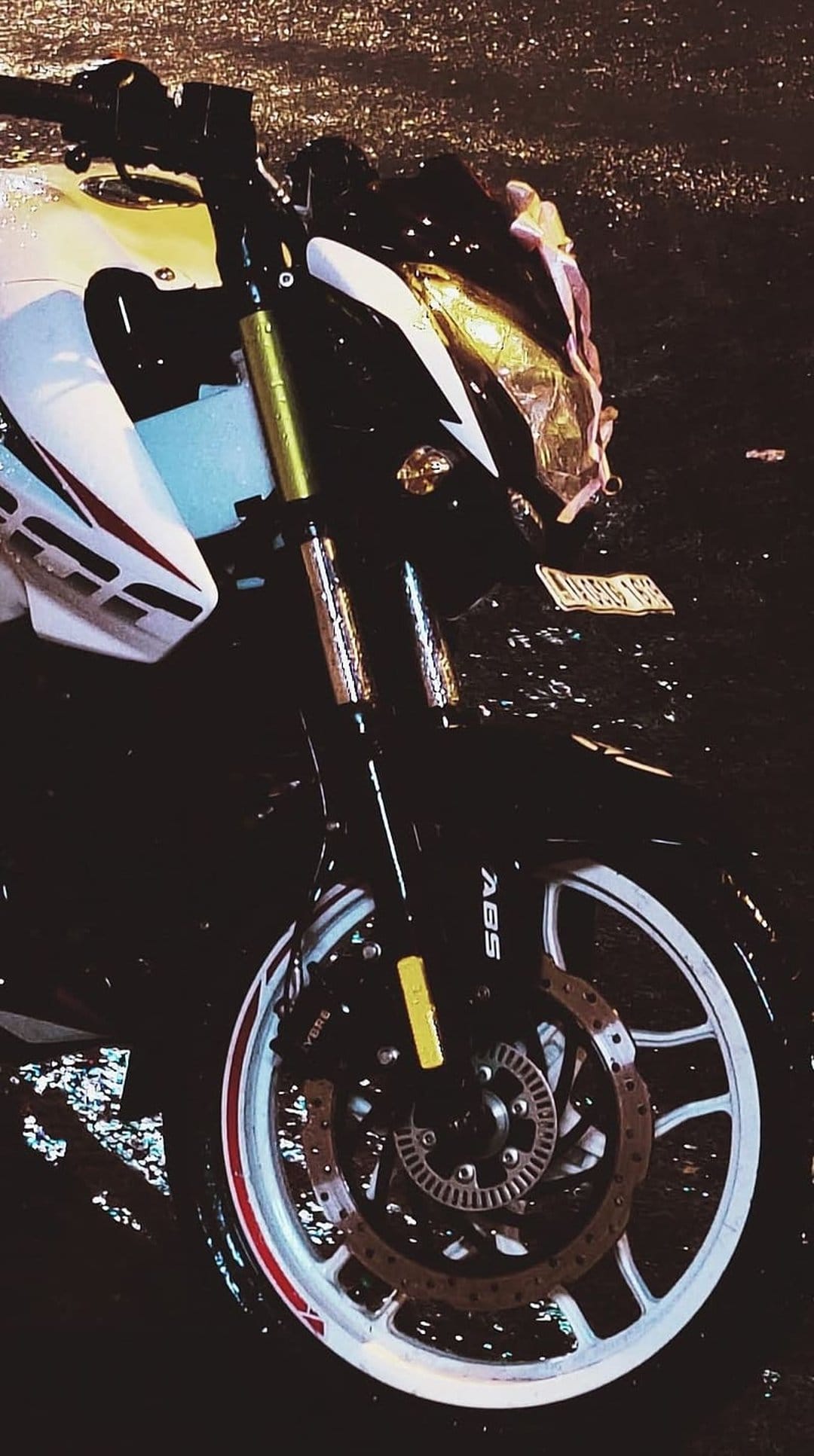 Motorcycle front wheel and ABS disc brake covered in raindrops with reflections on a wet asphalt street surface.
