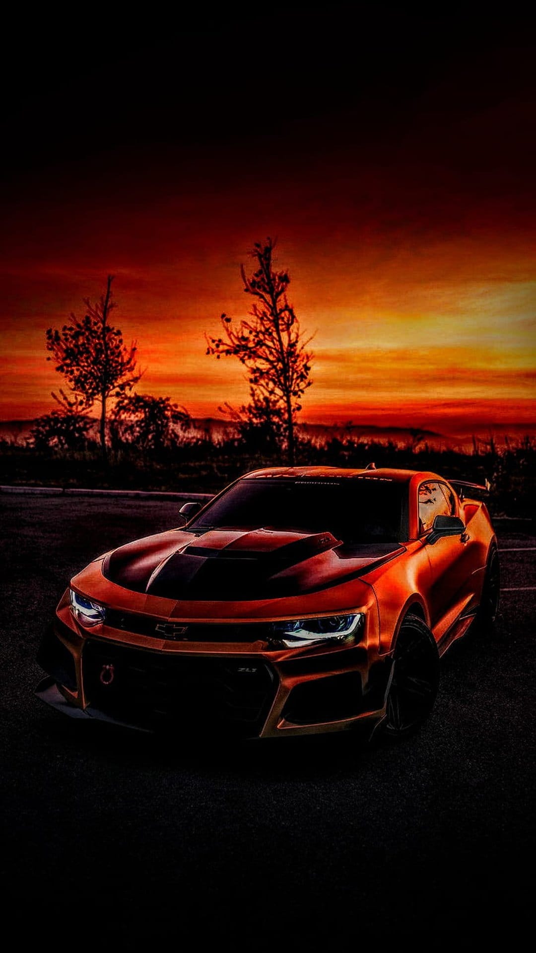 Orange Chevrolet Camaro parked on a road against a fiery red and orange sunset sky with dark tree silhouettes.