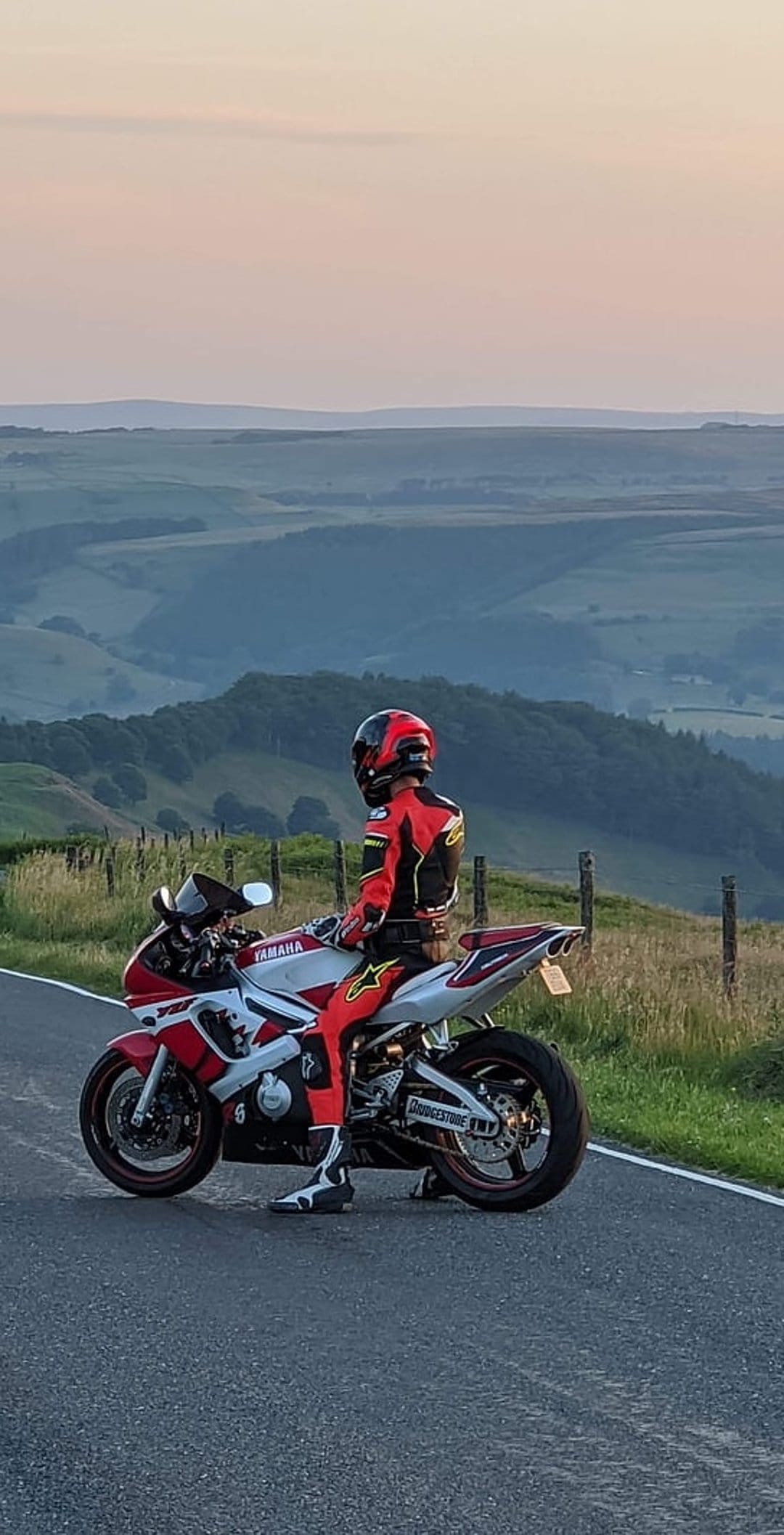 Motorcyclist in red racing gear on a Yamaha bike overlooking rolling green hills under a pink twilight sky.