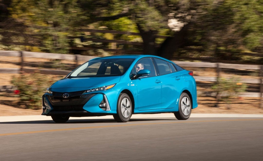 Blue Toyota Prius Prime driving on a sunlit road with blurred trees and a wooden fence in the background.